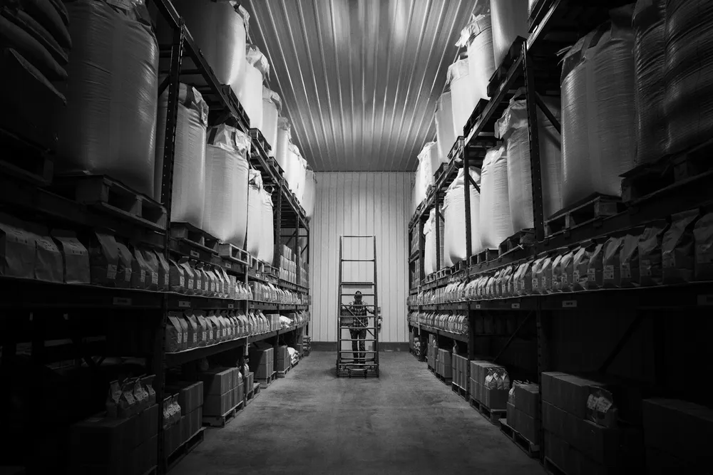 Black-and-white view down a warehouse aisle with tall racking holding sacks and boxed goods